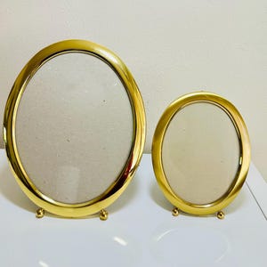 May include: Two gold-colored oval picture frames. The frames have a shiny finish and are supported by small, rounded feet. The oval openings are empty, ready for photos or artwork. The frames are different sizes.