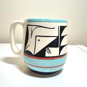 May include: A ceramic mug with a white handle and a cylindrical body. The mug features a geometric design in black, white, and blue. The rim and a band around the middle are light blue, with a red stripe. The base is dark blue.