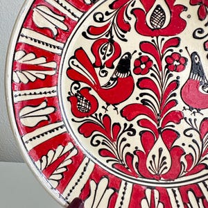 May include: A decorative ceramic plate with a cream-colored center and a red and black floral design. The rim features a red and white geometric pattern. Two red birds are featured in the center of the plate.