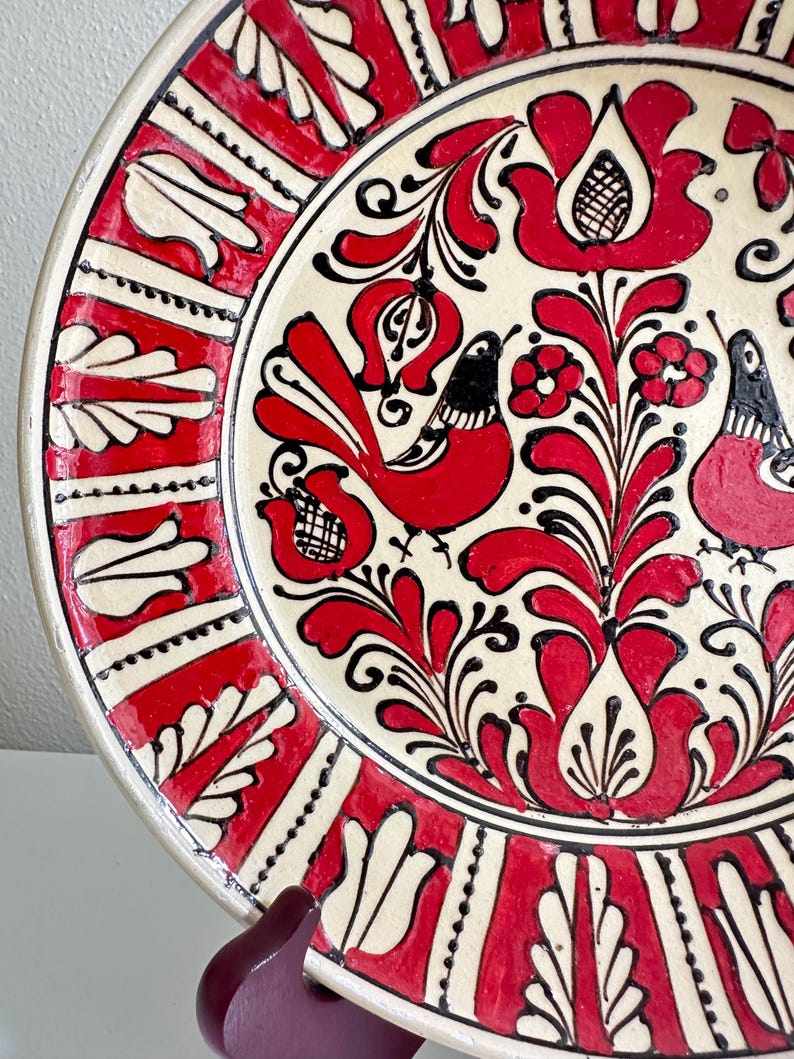 May include: A decorative ceramic plate with a cream-colored center and a red and black floral design. The rim features a red and white geometric pattern. Two red birds are featured in the center of the plate.