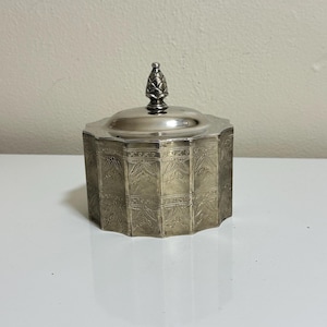 May include: A silver-toned metal lidded container with an octagonal shape and a pointed finial on top. The exterior features an intricate, repeating etched floral pattern across each panel, providing a textured appearance to the metallic surface.