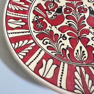May include: A decorative ceramic plate with a cream-colored base and a vibrant red and black floral design. The plate features stylized birds and intricate patterns, showcasing a traditional folk art style.