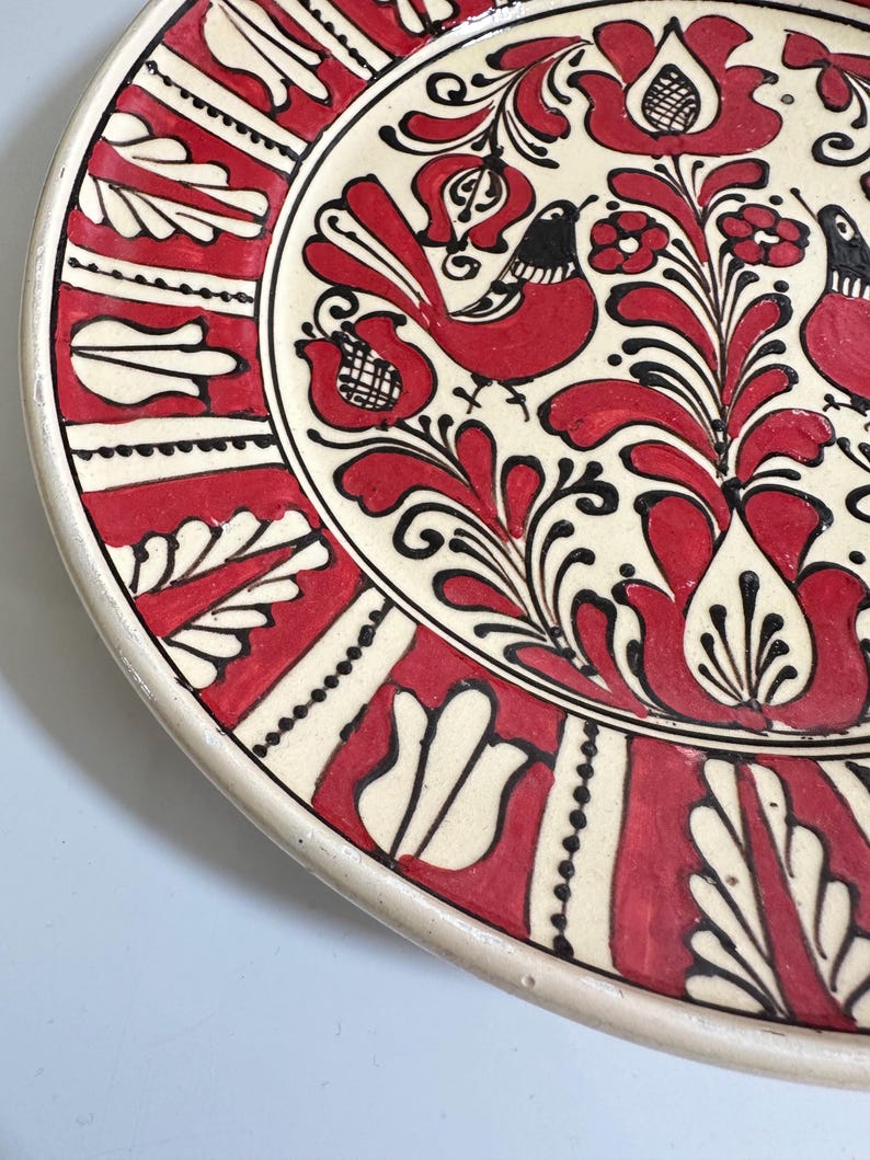 May include: A decorative ceramic plate with a cream-colored base and a vibrant red and black floral design. The plate features stylized birds and intricate patterns, showcasing a traditional folk art style.