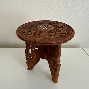 May include: A small, intricately carved wooden stool with a round top and three legs. The top features a floral design with a central inlaid star.