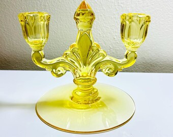 May include: A yellow glass candelabra with two candle holders and a central decorative element. The candelabra has a round base and is made of a transparent yellow glass. The design is ornate and vintage.