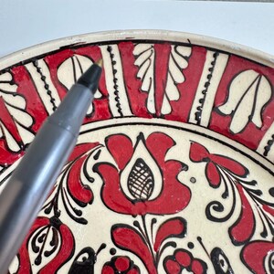 May include: A close-up of a decorative ceramic plate with a floral design. The plate features a cream-colored base with red and black painted floral patterns. The design includes stylized flowers, leaves, and other organic shapes.