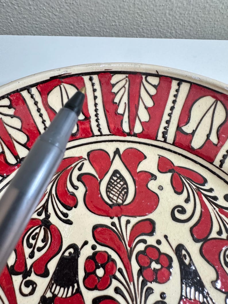 May include: A close-up of a decorative ceramic plate with a floral design. The plate features a cream-colored base with red and black painted floral patterns. The design includes stylized flowers, leaves, and other organic shapes.