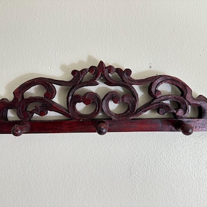 May include: A dark reddish-brown wooden wall-mounted coat rack featuring an intricate, symmetrical carved scrollwork design along the top edge. The horizontal base includes three rounded wooden pegs extending outward for hanging items.