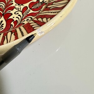 May include: A portion of a decorative ceramic plate with a cream-colored rim and a vibrant red and black floral and bird design. The plate's design is hand-painted, showcasing intricate details and a folk art style. A black pen is in the foreground.