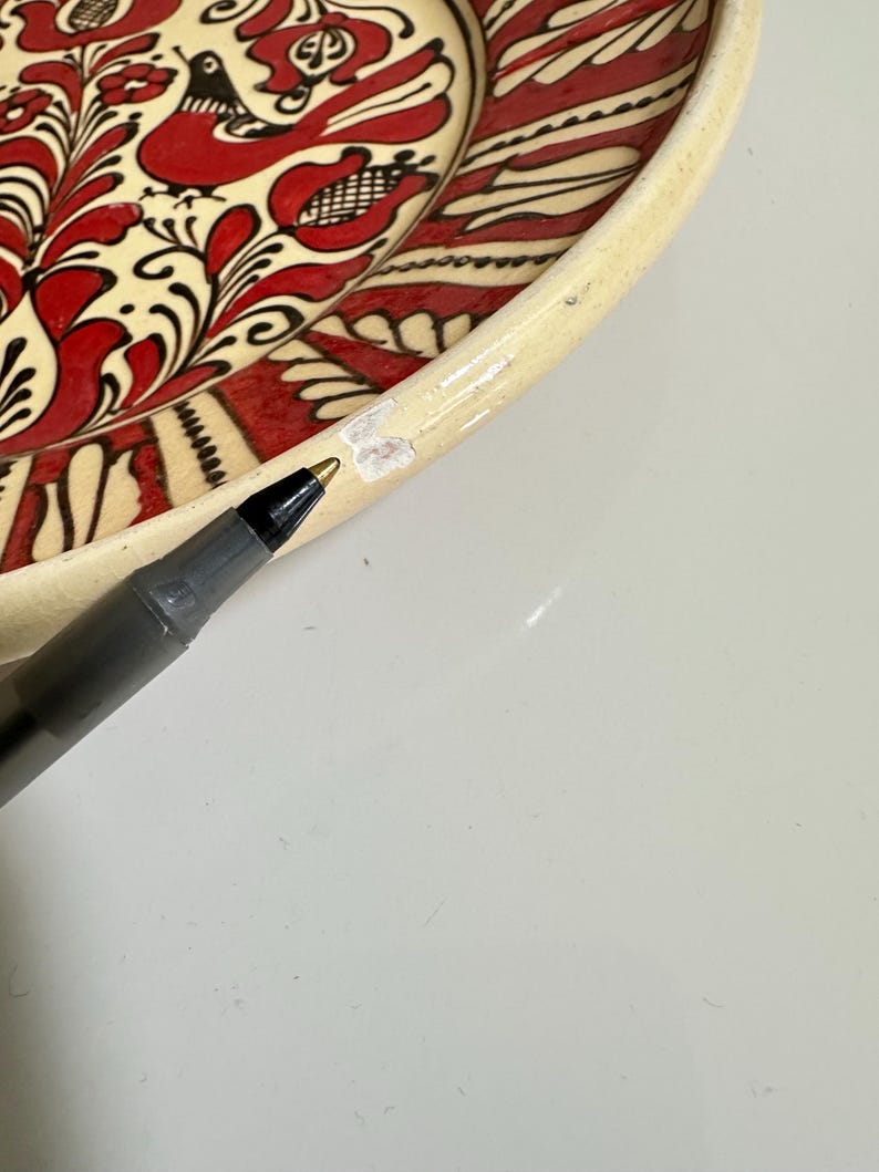 May include: A portion of a decorative ceramic plate with a cream-colored rim and a vibrant red and black floral and bird design. The plate's design is hand-painted, showcasing intricate details and a folk art style. A black pen is in the foreground.