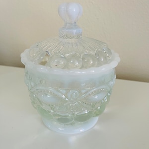 May include: A white milk glass candy dish with a clear glass lid and a heart-shaped knob. The dish has a scalloped edge and a floral design.