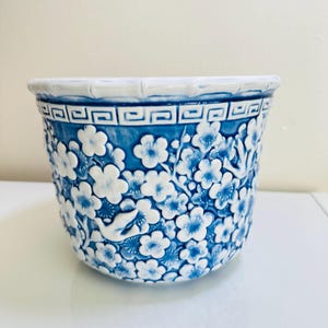 May include: A blue and white ceramic planter with a floral design. The planter features a repeating pattern of white flowers against a blue background. The rim has a geometric design. A bird is visible in the floral pattern.