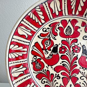 May include: A ceramic plate with a vibrant red and black floral and bird design on a cream background. The rim features a repeating pattern of red and white shapes. The plate's design is a folk art style.
