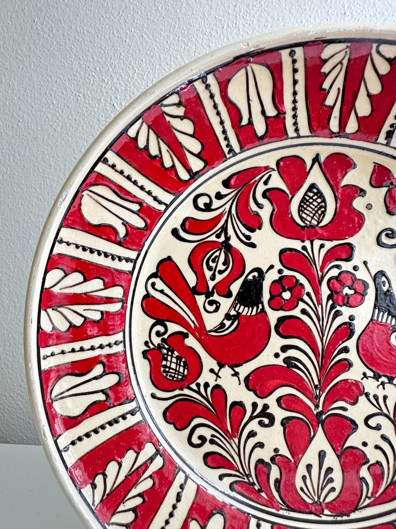 May include: A ceramic plate with a vibrant red and black floral and bird design on a cream background. The rim features a repeating pattern of red and white shapes. The plate's design is a folk art style.
