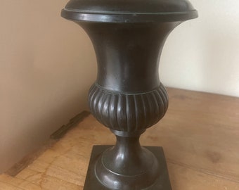 May include: A small, black, decorative urn with a fluted body and a square base.