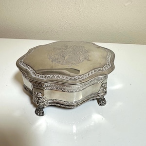 May include: A silver-toned metal box with a scalloped edge and a hinged lid. The lid features an engraved crest with a lion motif. The box has a decorative border and is supported by four ornate feet shaped like animal paws with detailed fur.