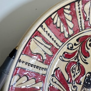 May include: Close-up of a decorative ceramic plate with a cream-colored base and intricate red and black floral patterns. The plate's rim features a repeating design. A black pen is in the frame.