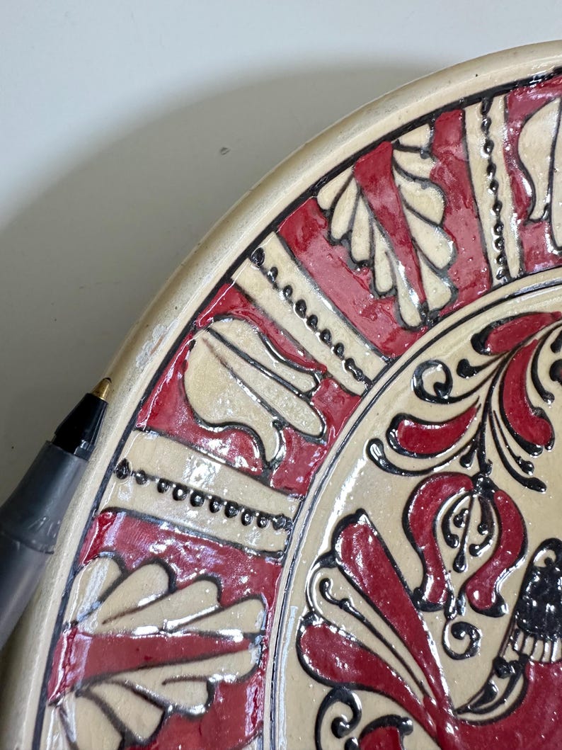 May include: Close-up of a decorative ceramic plate with a cream-colored base and intricate red and black floral patterns. The plate's rim features a repeating design. A black pen is in the frame.