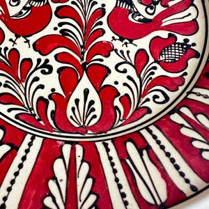 May include: A decorative ceramic plate with a hand-painted design. The plate features a white background with red floral and bird motifs, outlined in black. The rim has a repeating pattern of red and white vertical stripes. A traditional folk art piece.