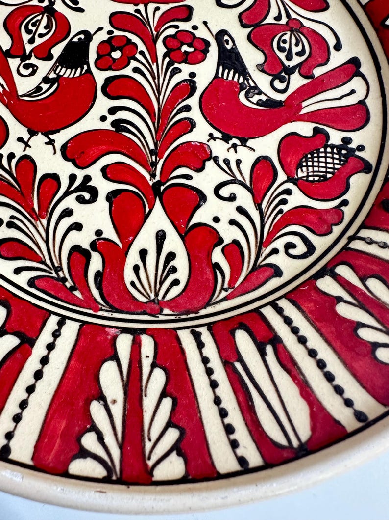May include: A decorative ceramic plate with a hand-painted design. The plate features a white background with red floral and bird motifs, outlined in black. The rim has a repeating pattern of red and white vertical stripes. A traditional folk art piece.