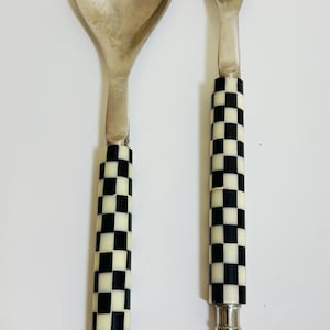 May include: A silver salad spoon and fork set. The spoon has a rounded bowl, and the fork has three tines. Both utensils have black and white checkered handles. The set is designed for serving salads and other dishes.
