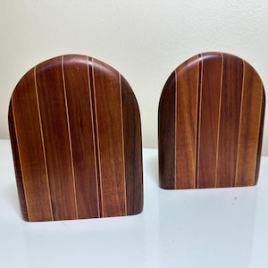 May include: A pair of arched bookends crafted from vertical strips of dark and medium-toned wood. Each piece features a smooth, rounded top edge and a flat base, showcasing the natural grain patterns and alternating color variations of the wood.