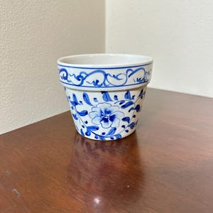 May include: A white ceramic flower pot featuring hand-painted blue floral patterns and scrolling vine motifs around the exterior. The rim is decorated with a continuous blue line design, and the base tapers slightly toward the bottom.
