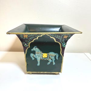 May include: A dark green metal planter with a square top and a decorative horse painting. The planter has a gold trim and is painted with a horse design in the center. The planter is approximately 6 inches tall.