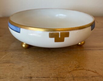 May include: A white ceramic bowl with a gold rim and blue and gold geometric designs. The bowl has three gold feet.