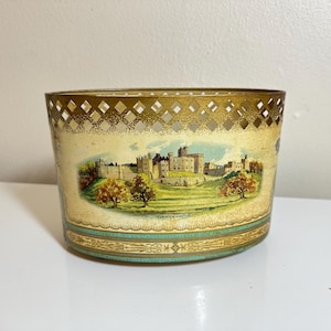 May include: An oval metal container featuring a cream-colored body with a detailed illustration of Alnwick Castle set in a green landscape. The top rim has a gold-toned lattice cutout design, and the base is decorated with teal and gold trim.