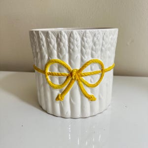 Peut inclure: Un cache-pot en céramique blanche présentant une texture moulée en forme de tiges d'asperges sur l'extérieur. Un détail jaune vif rappelant une corde entoure le centre du pot, noué en un nœud décoratif sur le devant du récipient cylindrique.