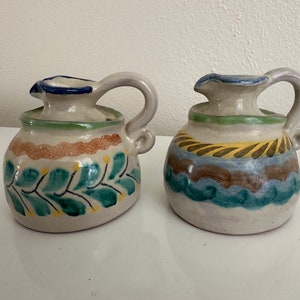 May include: Two small ceramic pitchers with colorful painted designs. One pitcher has a green and yellow floral pattern, while the other has a blue, yellow, and brown geometric pattern.