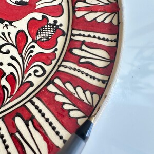 May include: Close-up of a decorative ceramic plate with a traditional folk art design. The plate features a cream-colored base with red and black floral and geometric patterns. The design includes stylized flowers and leaf motifs.