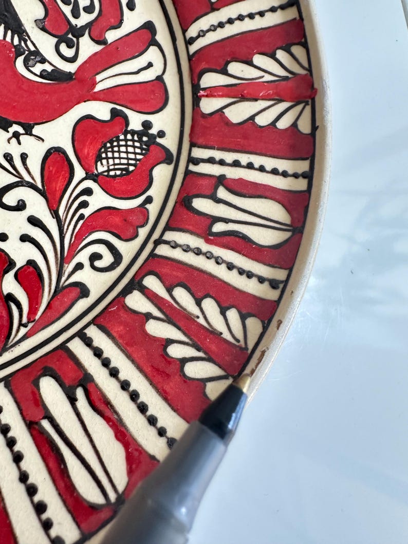May include: Close-up of a decorative ceramic plate with a traditional folk art design. The plate features a cream-colored base with red and black floral and geometric patterns. The design includes stylized flowers and leaf motifs.