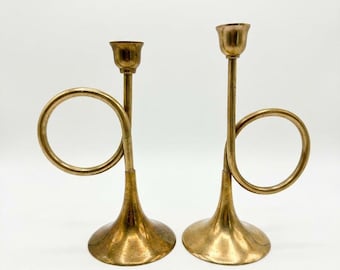 May include: A pair of brass candle holders shaped like post horns. Each holder features a circular loop, a slender vertical stem, a flared base, and a small cup at the top designed to hold a single taper candle. The metal has a polished finish.