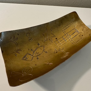 May include: A gold-colored metal tray with a curved edge and an etched design of geometric shapes and symbols. The tray is approximately 12 inches long.