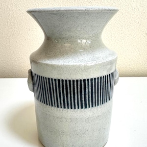 May include: A speckled, light gray ceramic vase with a flared top and a cylindrical body. The vase features a band of vertical dark blue stripes around the middle and small loop handles. A decorative piece for the home.