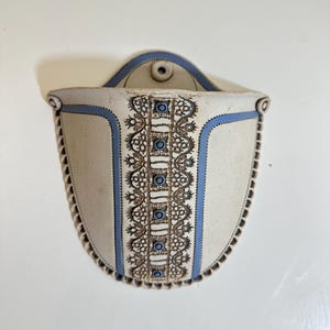 May include: A wall-mounted ceramic pocket planter in a matte cream finish with a blue border. The front features a vertical brown lace-like pattern with small blue circular accents. It has a semi-circular shape with a scalloped edge and a single hole.