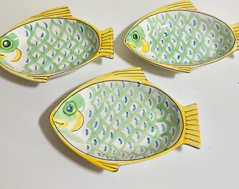 May include: Three ceramic dishes shaped like fish, each featuring a yellow border, a green and white scale pattern on the body, and a painted eye. The dishes have a concave center and are arranged in a triangular formation on a plain surface.
