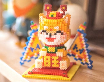 May include: A colorful beaded figurine of a dog wearing a red and yellow crown and a green and red robe. The dog is sitting on a red and yellow beaded mat. The figurine is made of small plastic beads.