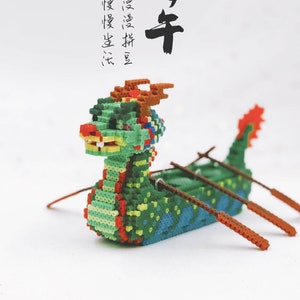 May include: A green, red, and yellow dragon boat made of small plastic beads. The boat has four brown oars and the text "端午节快乐" is written in black ink above the boat.