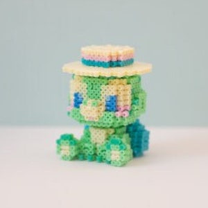 Puede incluir: Una figura de tortuga pixelada en tonos verdes, azules y amarillos, con un pequeño sombrero de paja con una banda rosa y azul. La tortuga está sentada sobre una superficie blanca contra un fondo azul claro.