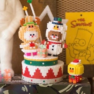 May include: A festive Christmas-themed display featuring pixelated figures. A reindeer and snowman sit atop a tiered cake, with a small chick and gift boxes nearby. A white astronaut statue and a yellow "The Simpsons" sign are in the background.
