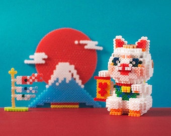 May include: A white bead figurine of a Japanese Maneki-Neko, or beckoning cat, holding a red scroll. The cat is sitting on a red surface in front of a blue and white bead model of Mount Fuji, a red sun, and a white and blue bead model of a Japanese carp streamer.