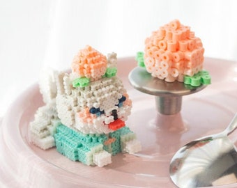 May include: A pink ceramic pot with a white lid. The lid has a small, white and green beaded figure of a cartoon animal with a pink flower on its head. A pink beaded flower is on the knob of the lid.