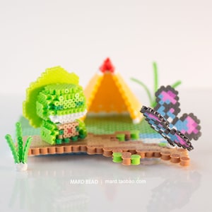 Könnte beinhalten: Eine grüne und weiße Perlen-Sprite-Figur mit einem gelben Hut steht vor einem gelben Zelt. Ein blauer und rosa Schmetterlings-Sprite aus Perlen fliegt über einer grünen Perlenpflanze. Die Perlen-Sprites befinden sich auf einer braunen Basis mit grünen Perlenakzenten.