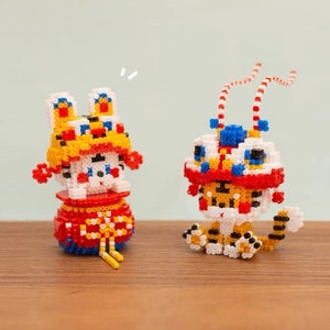 Könnte beinhalten: Zwei kleine, bunte, perlenbesetzte Figuren von Löwentänzern. Eine ist rot, weiß und gelb mit einem rot-weißen Hut. Die andere ist rot, weiß und blau mit einem rot-weißen Hut. Beide haben schwarze Perlenaugen.