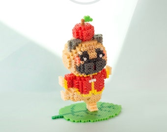 May include: A small, beige pug figurine wearing a red and yellow jacket and a red apple hat. The figurine is standing on a green leaf.