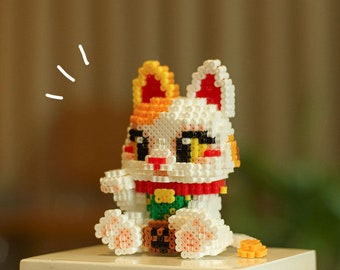 May include: A white, red, yellow, and green bead figurine of a Japanese Maneki-Neko, or lucky cat, with its paw raised. The figurine is sitting on a white surface.