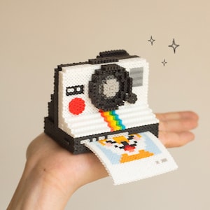 以下が含まれることがあります： 虹色のストライプと、カメラから出てくるピクセル化されたオレンジと白の動物の写真がついた、白と黒のピクセル化されたカメラです。